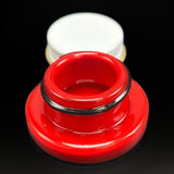 The cherry red glass jar open, the black band around its threaded neck, the lid behind it